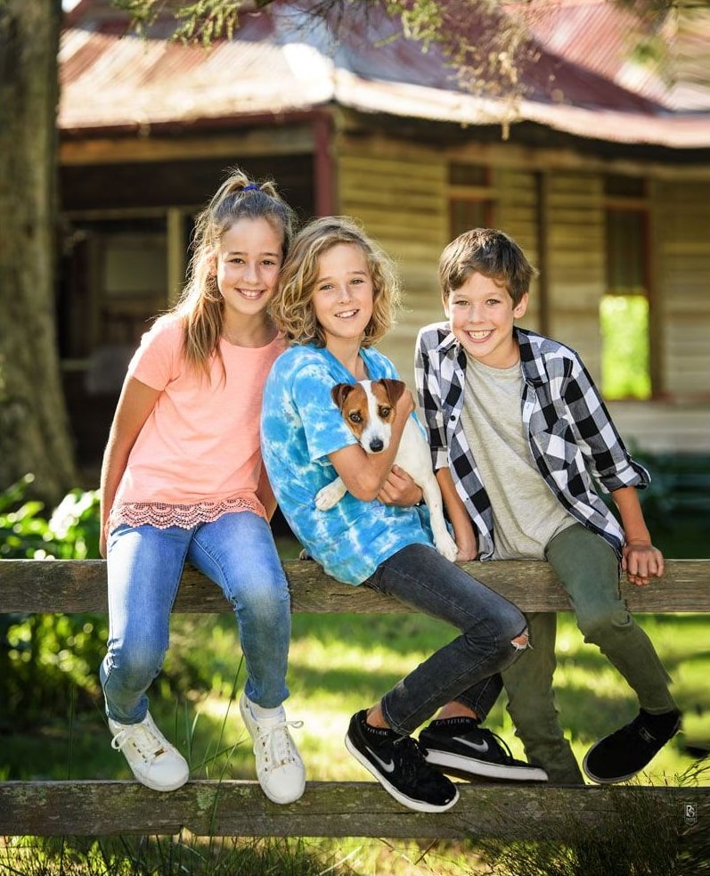 Three children on a timber farm fence, one cuddling a small dog, near an old cottage