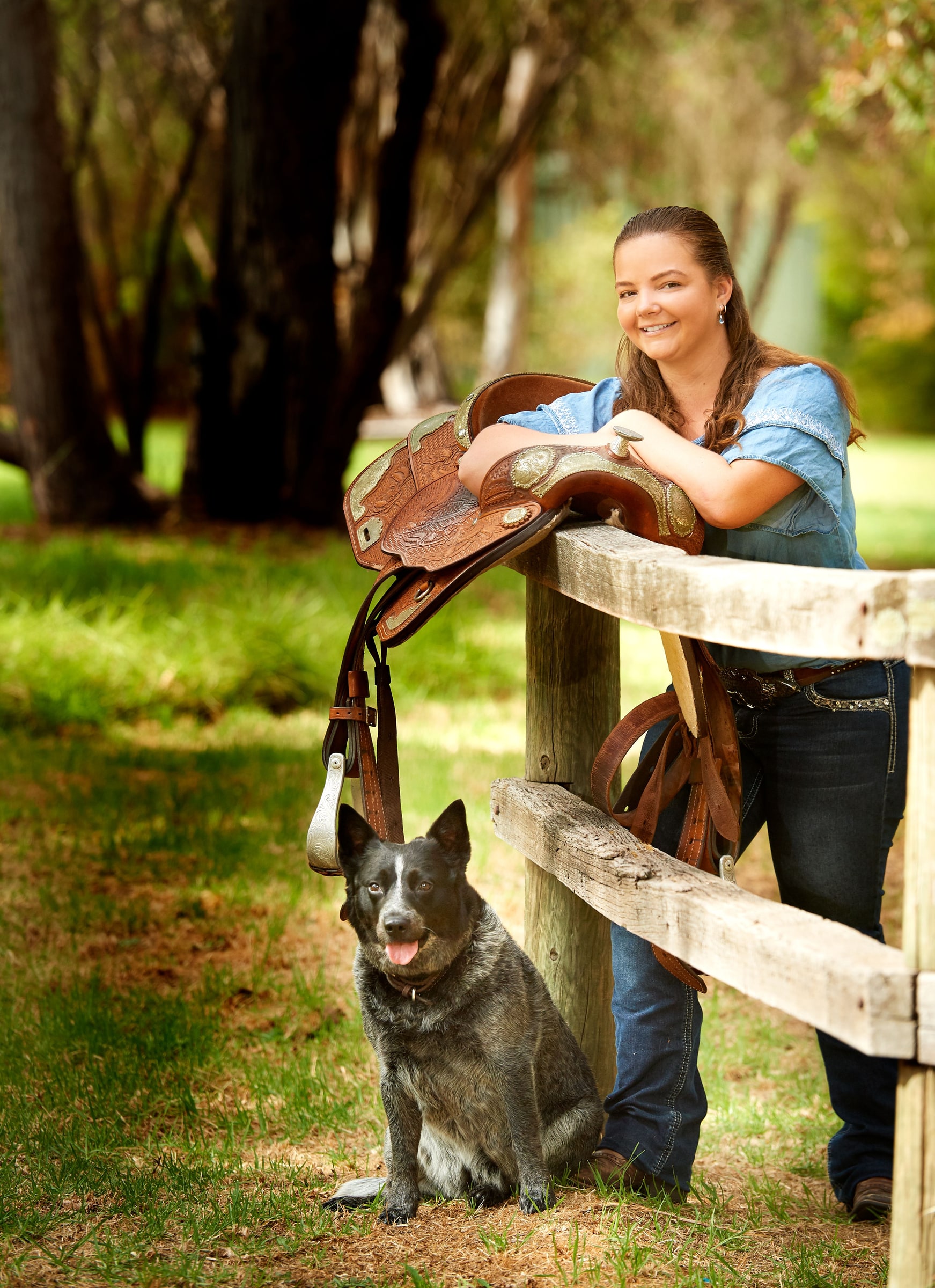A woman leaning on a fence hung with a saddle, her cattle dog at her feet