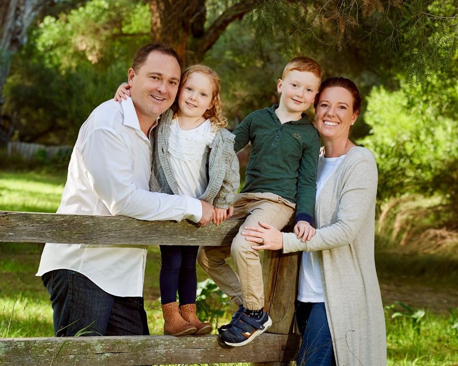 Parents hug a girl and boy perched on a timber farm fence among green trees