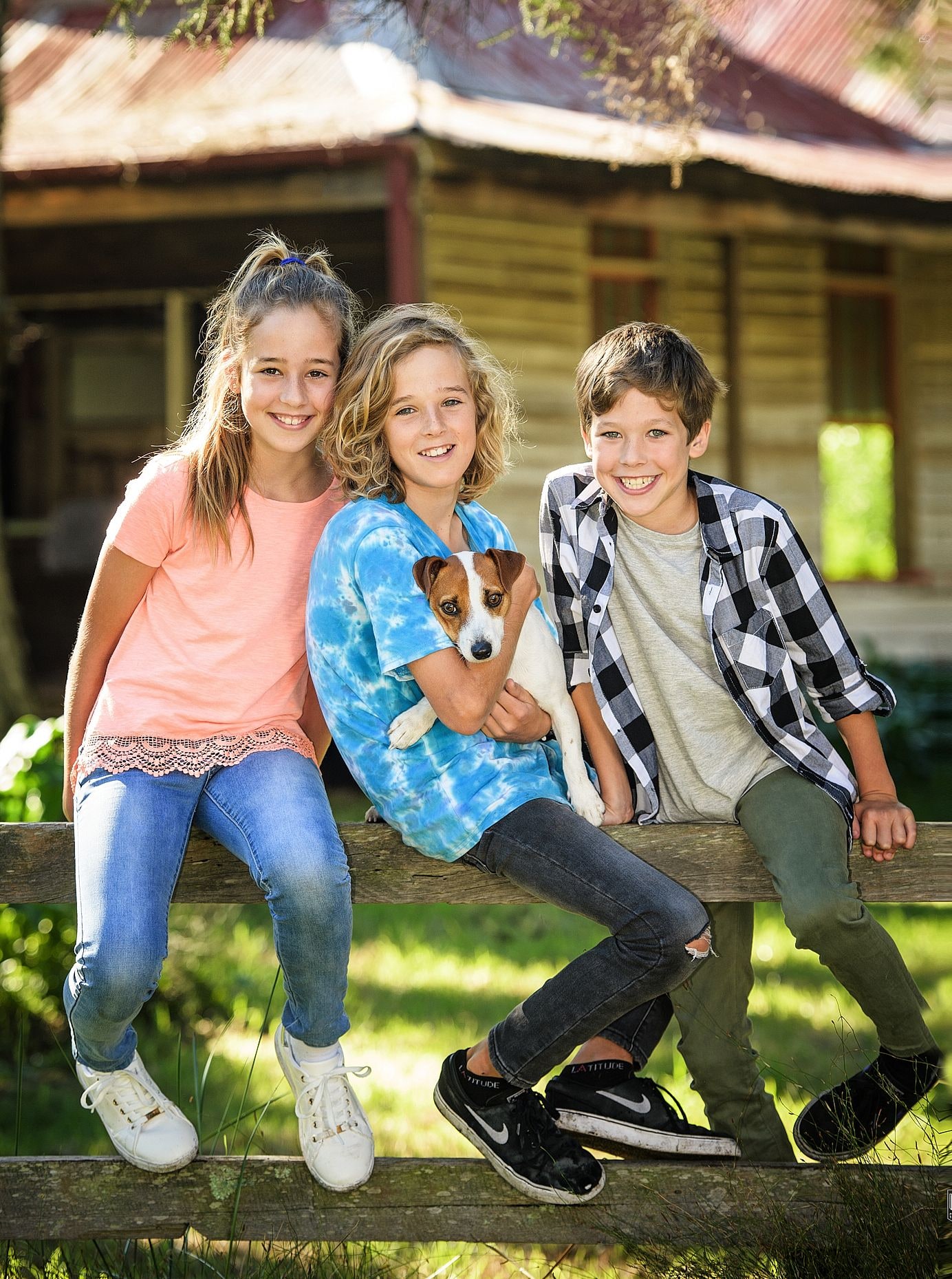 Three smiling children share a rustic fence rail with a small dog by a weatherboard cottage
