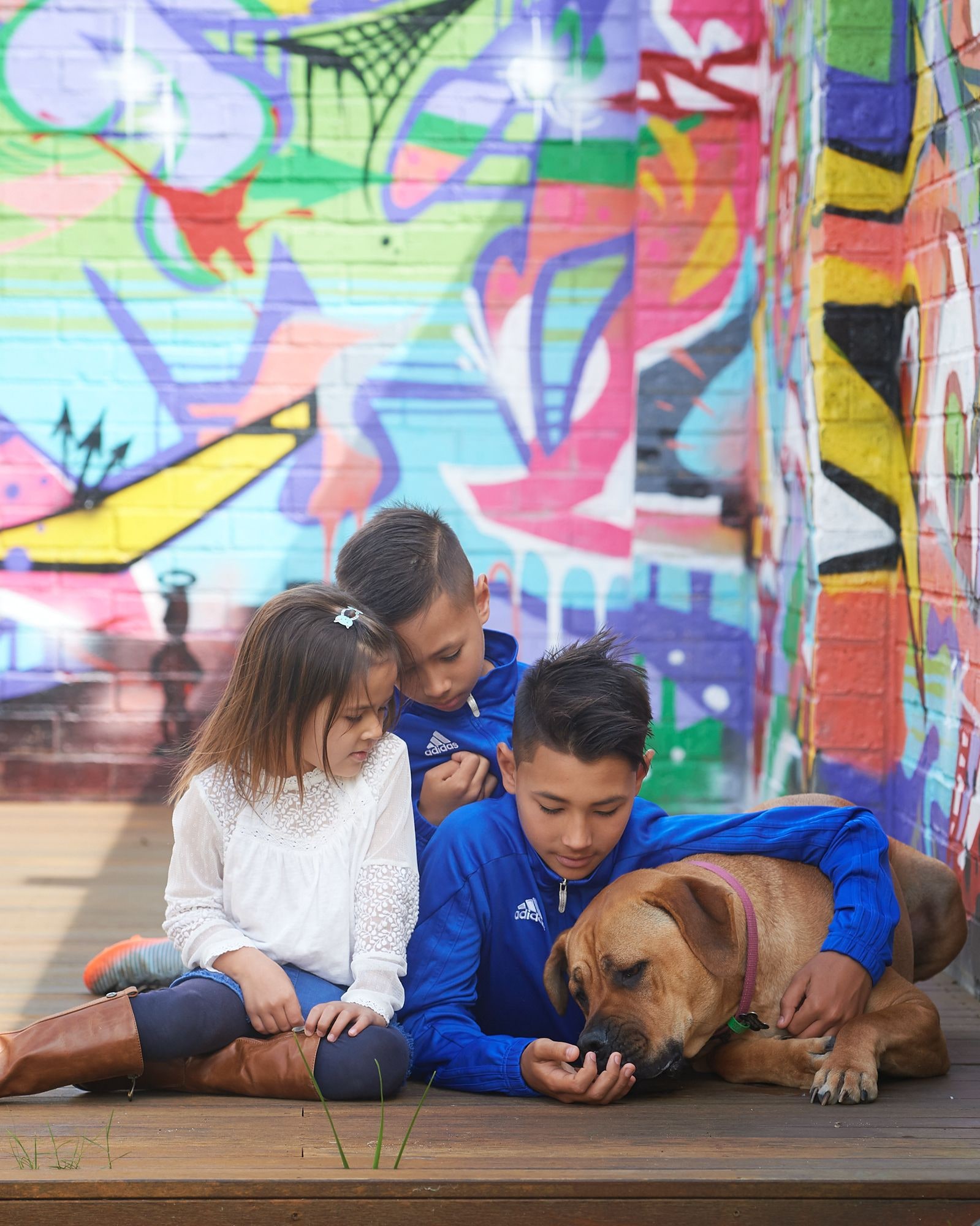 Three children cuddling their dog in front of the colourful graffiti