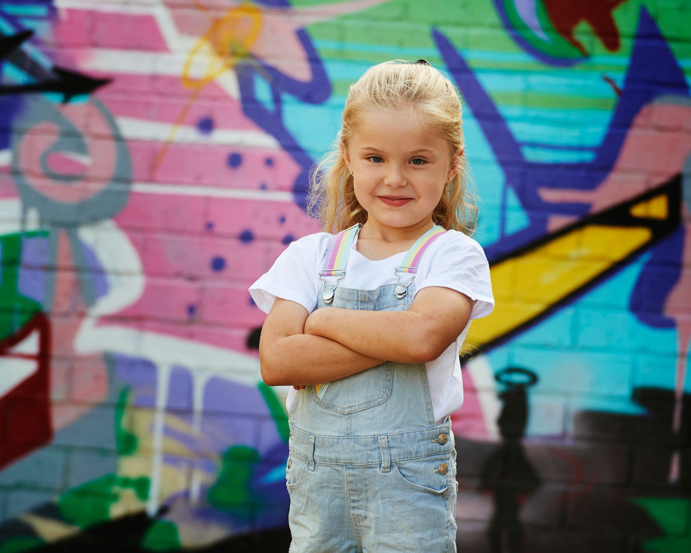 A little girl in overalls with her arms folded, in front of the graffiti