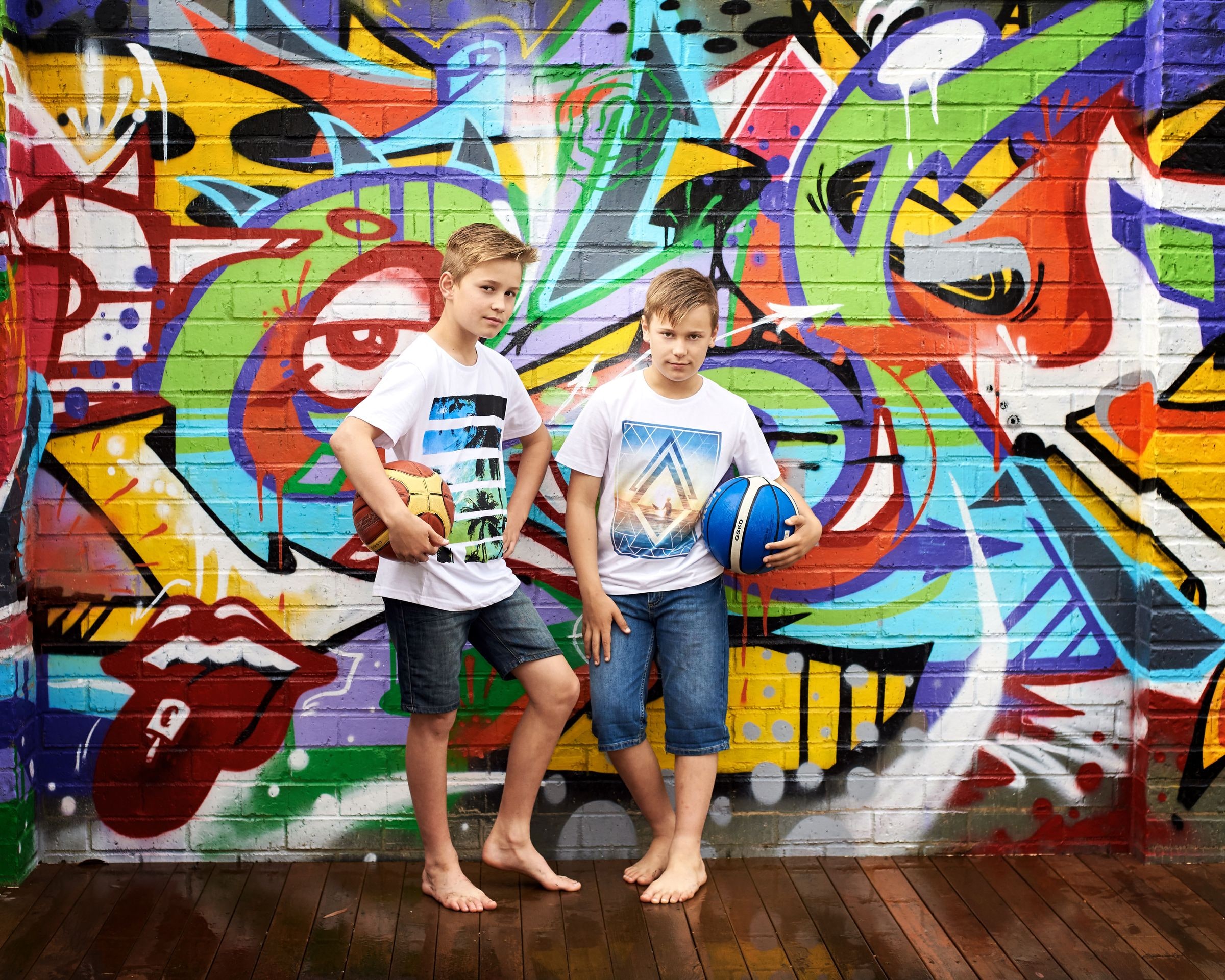 Two boys holding basketballs stand barefoot on a timber deck in front of bright graffiti