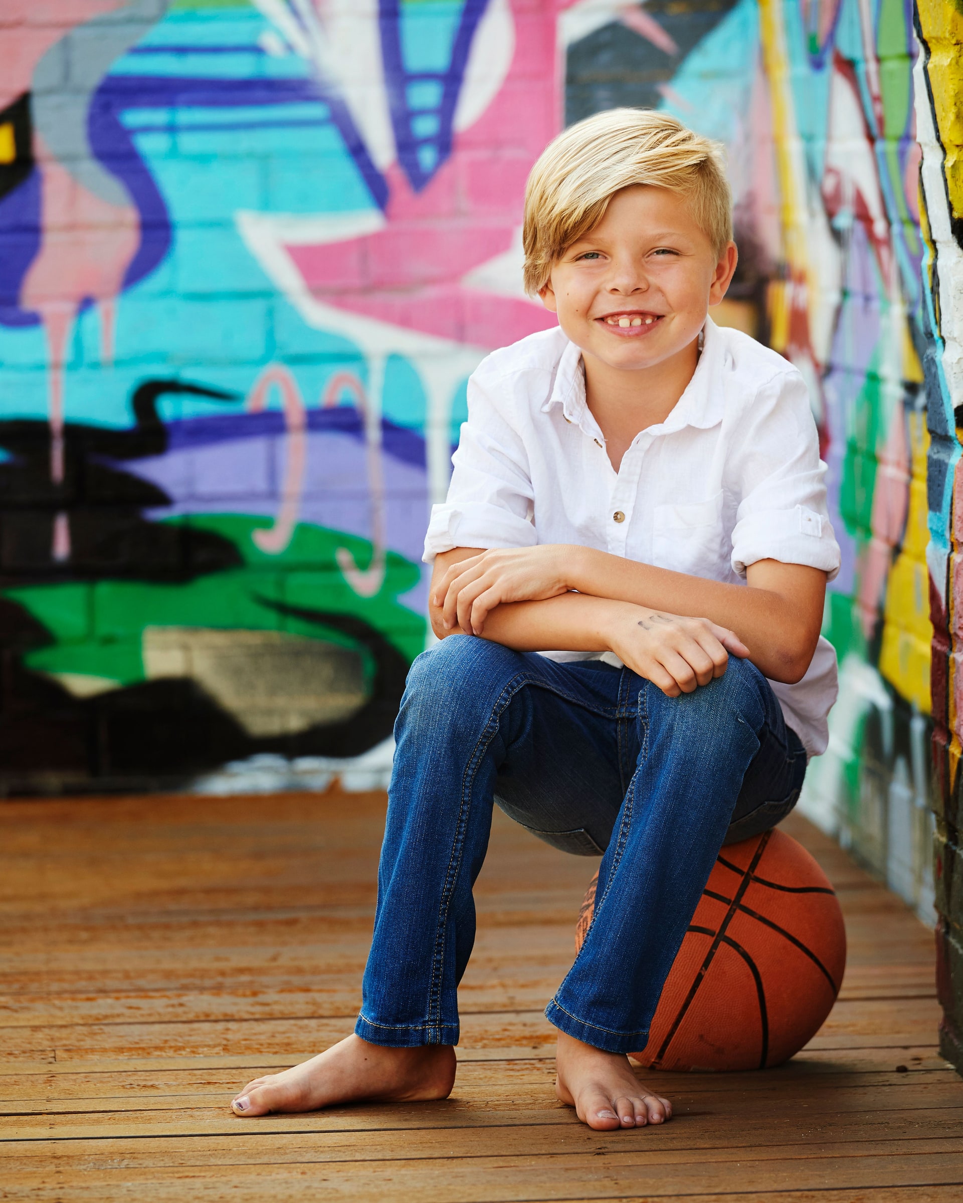 A boy sitting on a basketball against the graffiti wall