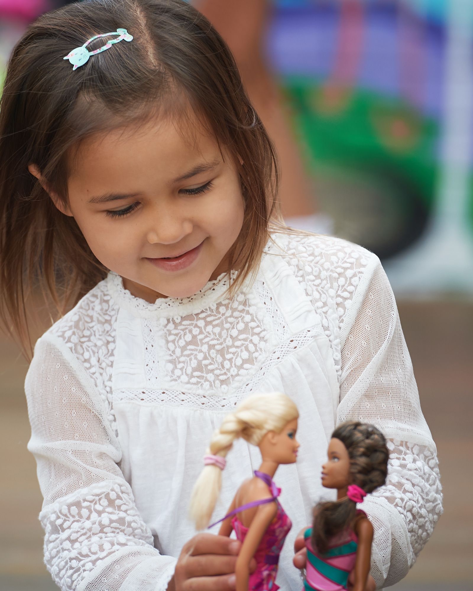 Young girl in a white lace top smiles at two dolls before a colourful graffiti wall