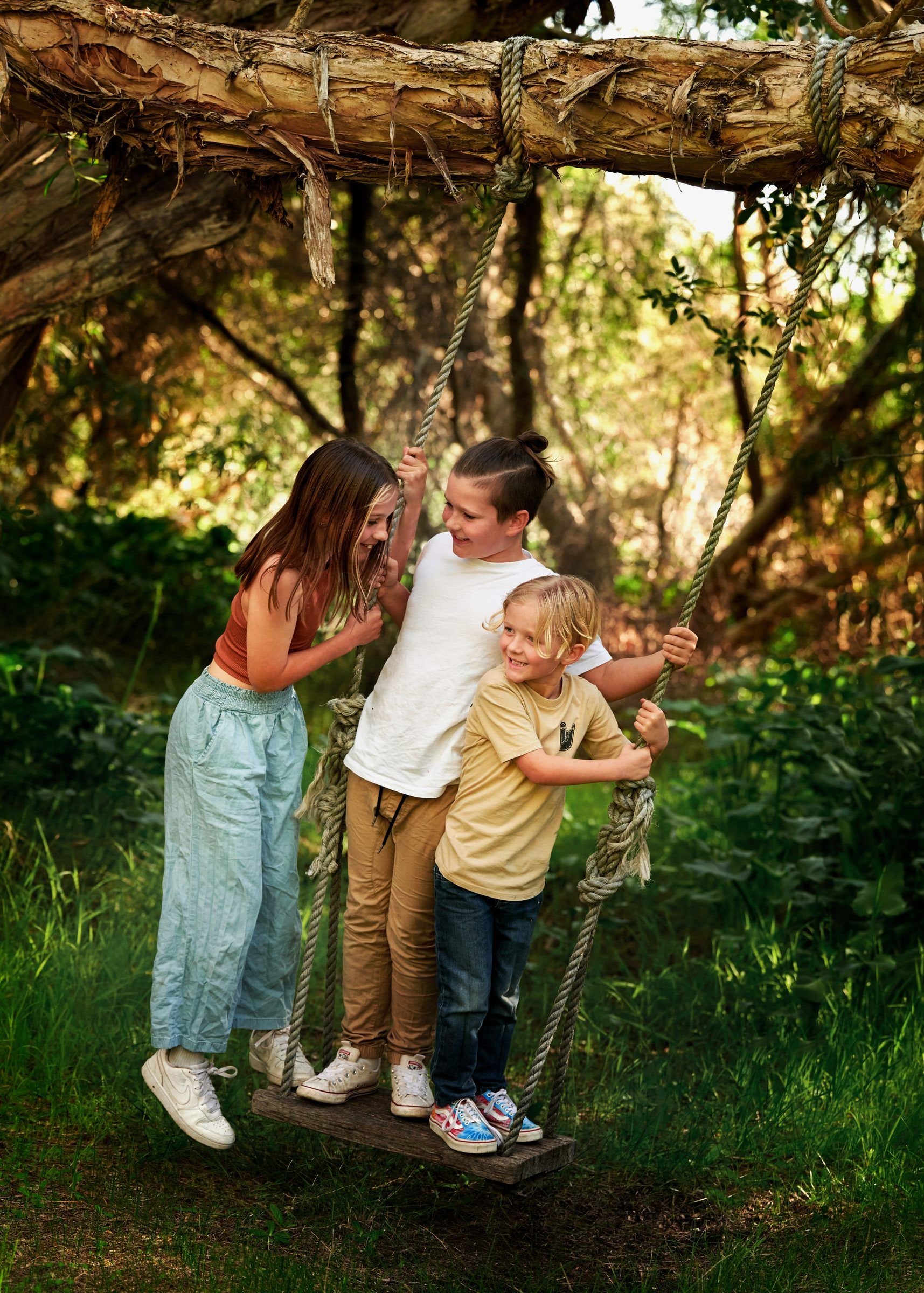 Three children stand together on a rope tree swing, smiling at each other among green bushland