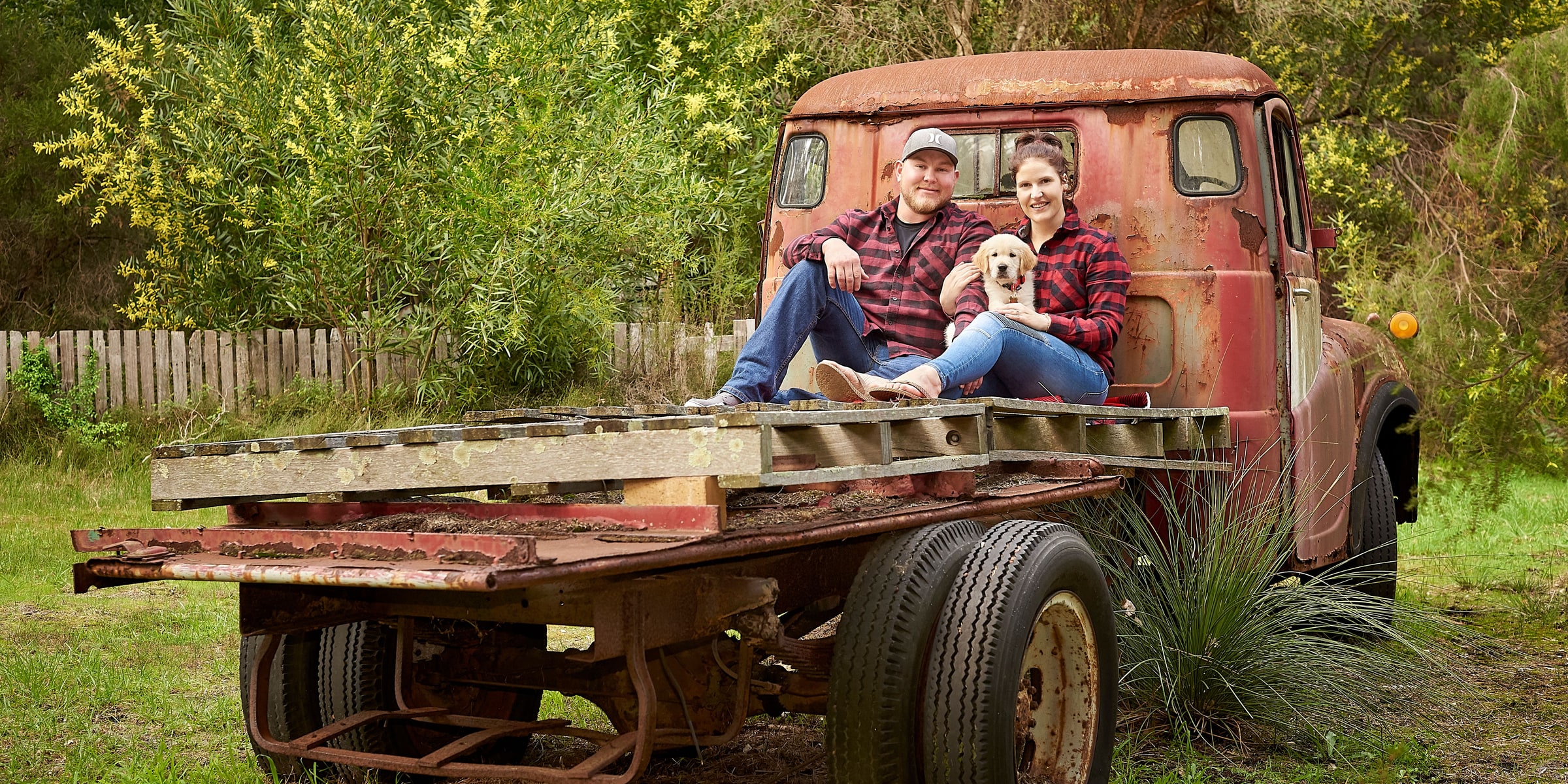 Couple in red flannel shirts sit on an old truck tray with a golden puppy