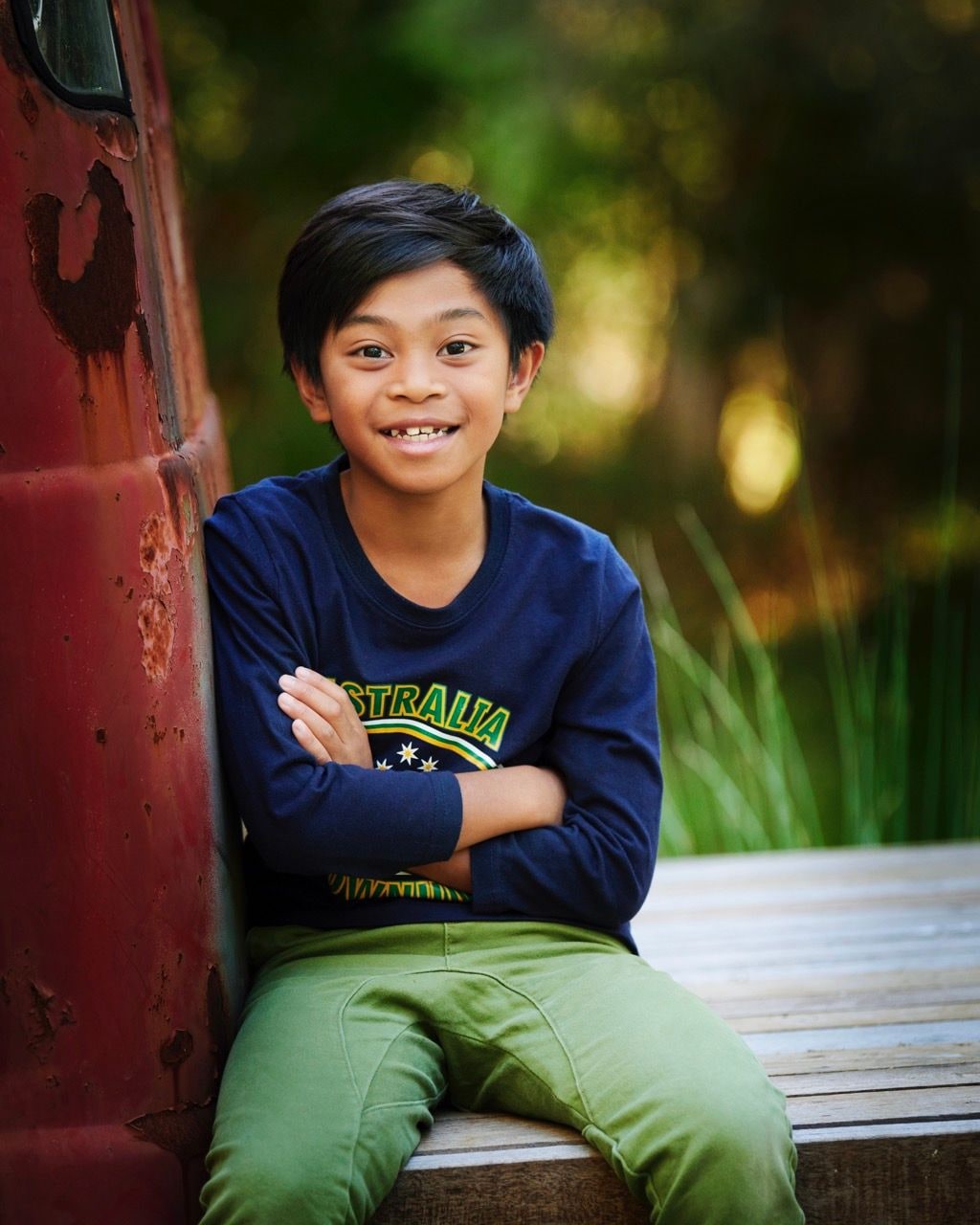 Smiling boy with folded arms leans against a rusty red truck on a timber deck
