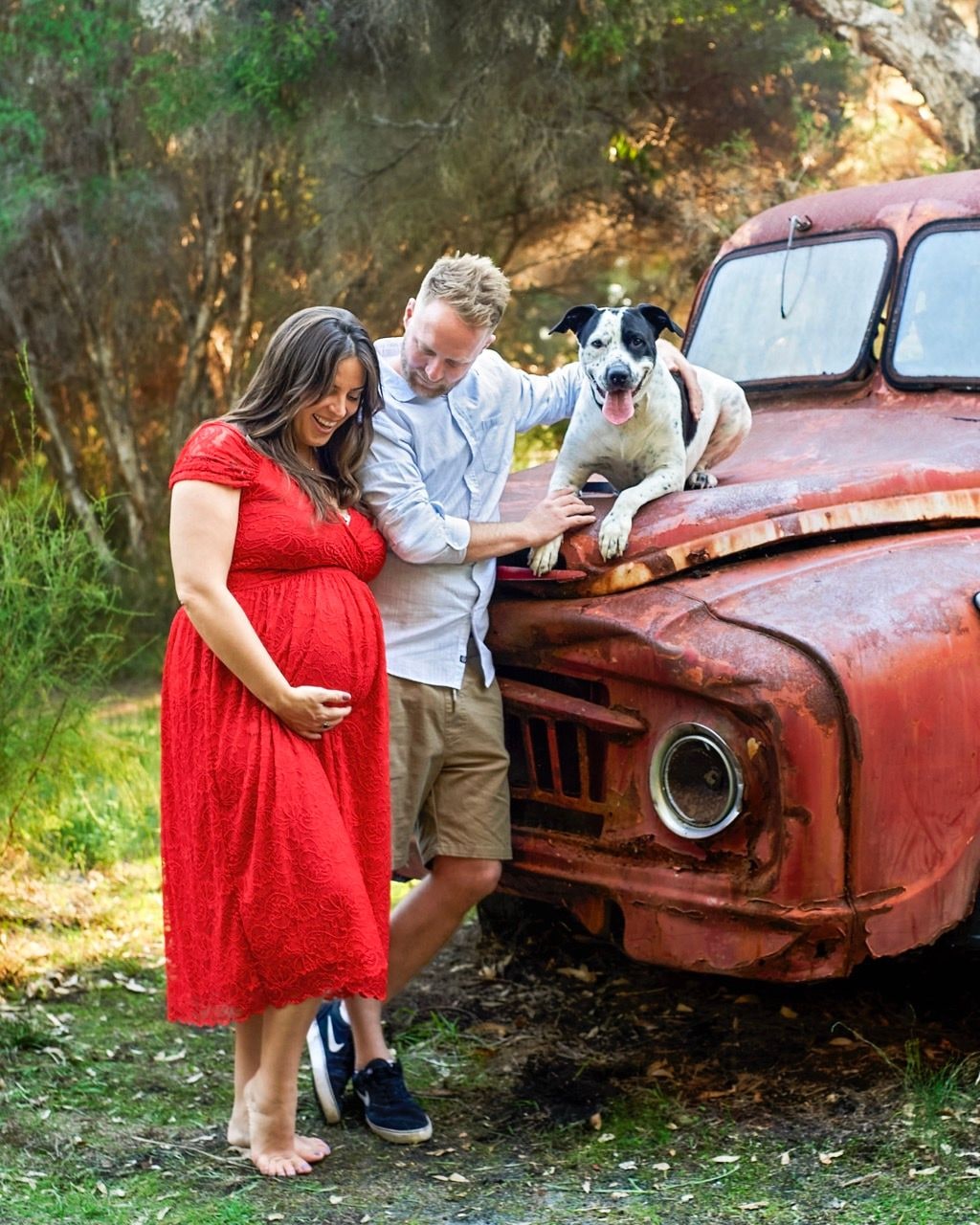Expectant couple stand beside a rusty red truck while their dog lies on the bonnet