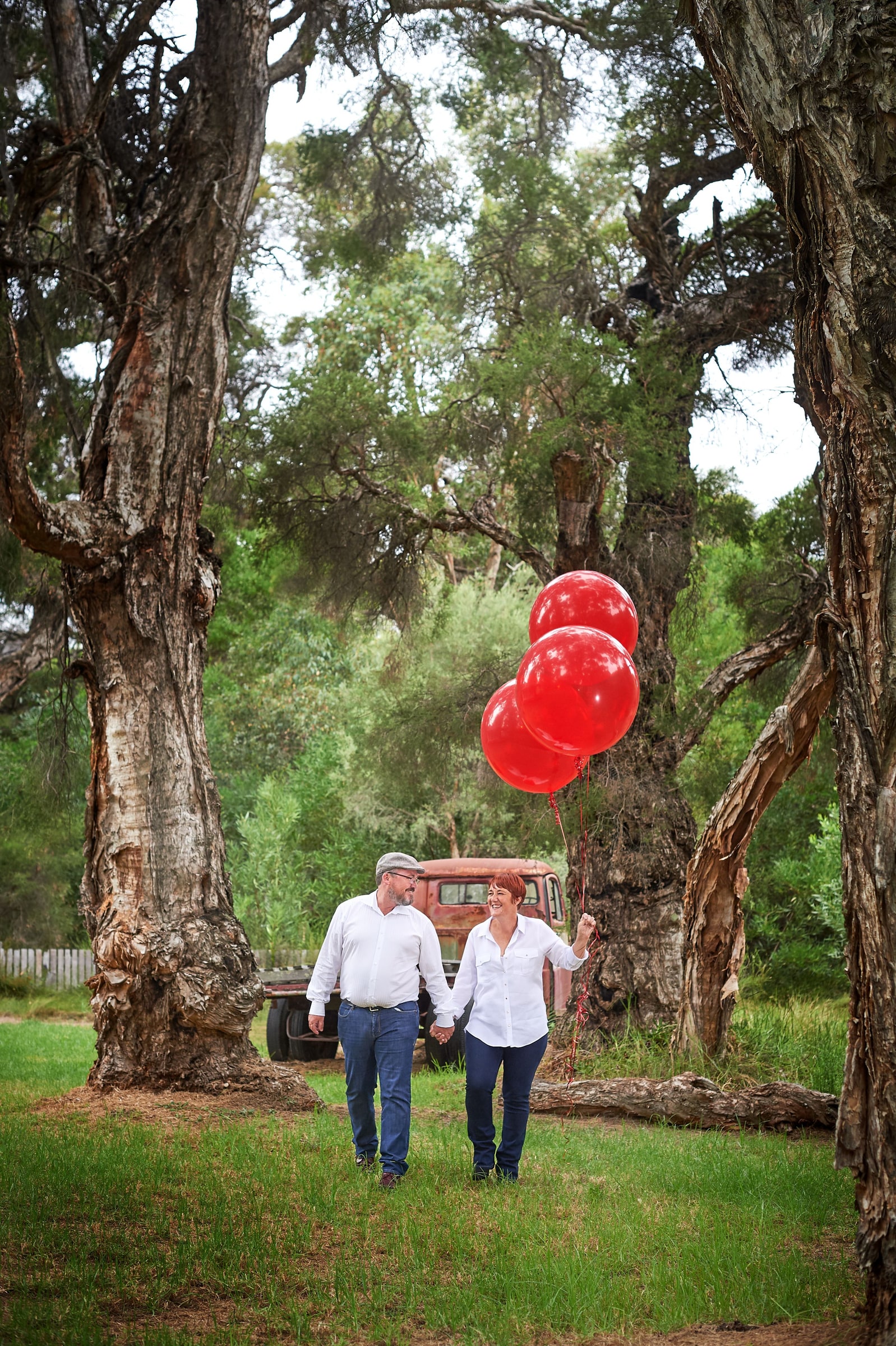 Couple walk hand in hand under tall paperbark trees holding three red balloons