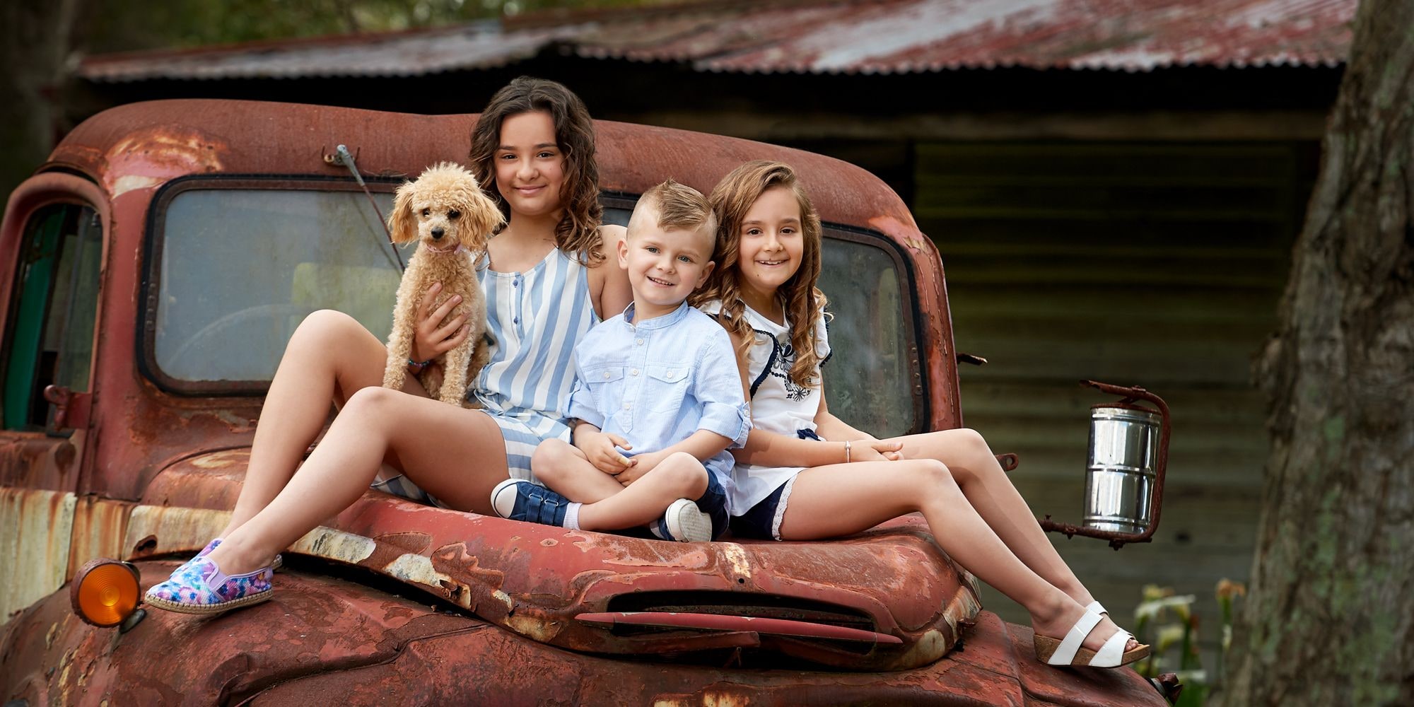 Three children and a fluffy poodle sit on the bonnet of a rusty vintage truck