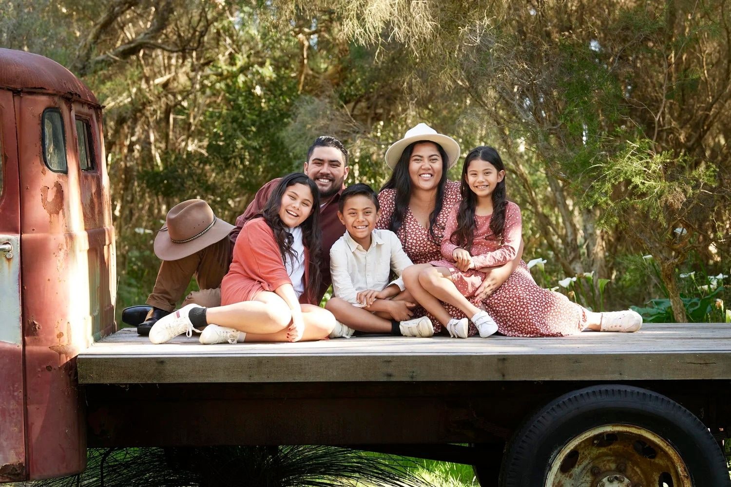 A family of six sitting on the tray of the vintage red truck among the trees