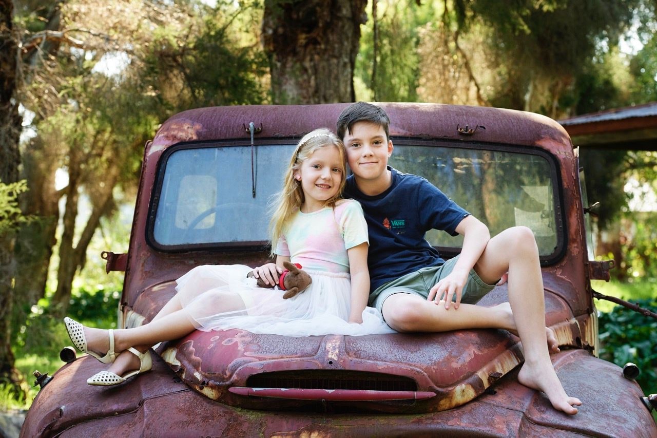 Young girl and boy sit together on the bonnet of a rusty vintage truck