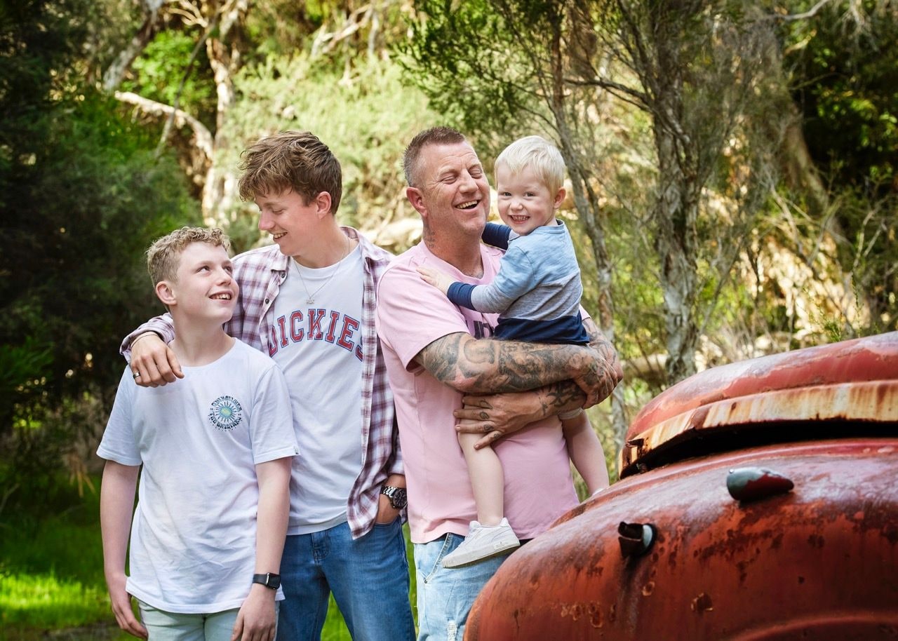Laughing man holds a toddler beside two older boys next to a rusty red truck bonnet