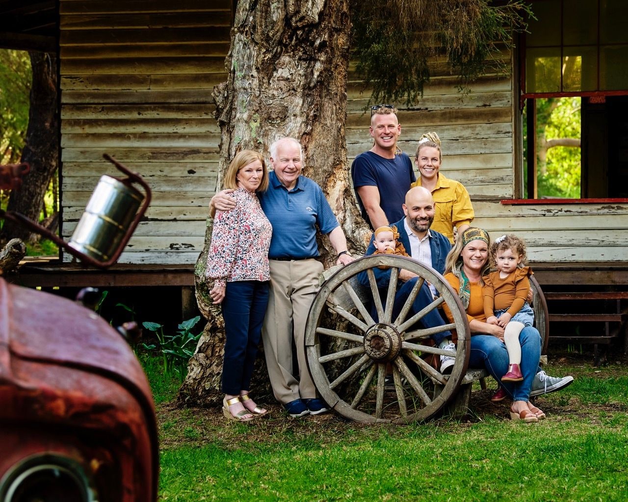 Three-generation family gathers around a wagon wheel in front of an old weatherboard cottage