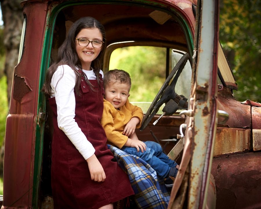 Girl in glasses and a little boy sit in the open cab of a rusty truck
