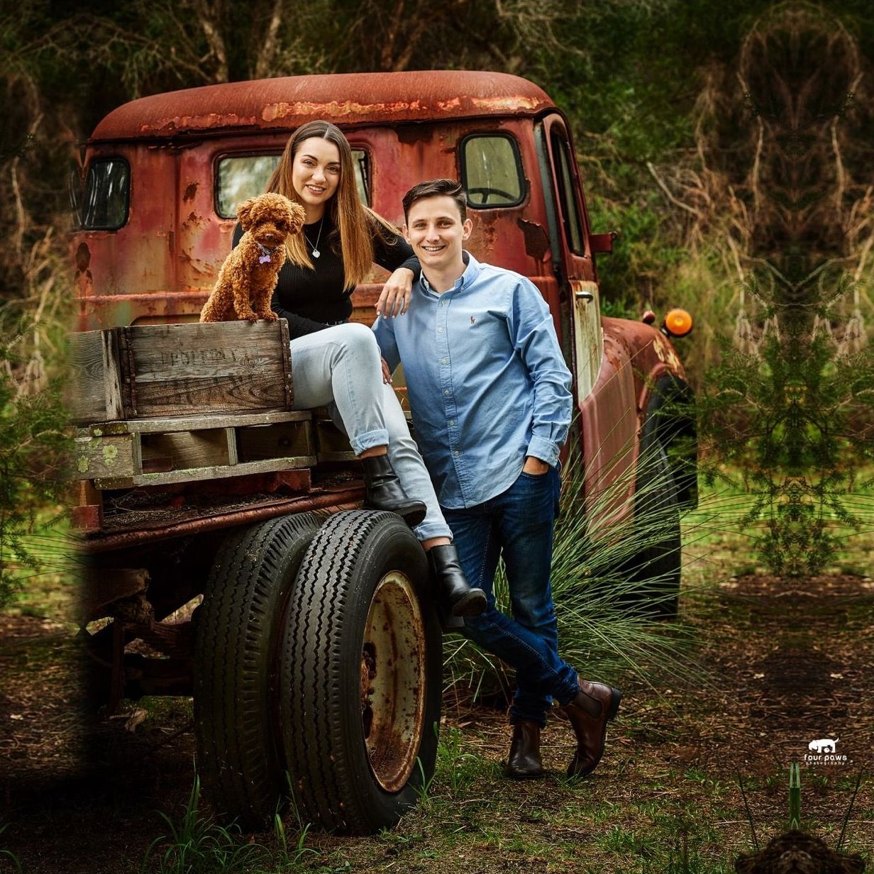 Woman holding a curly poodle sits on a rusty red truck tray beside a smiling man