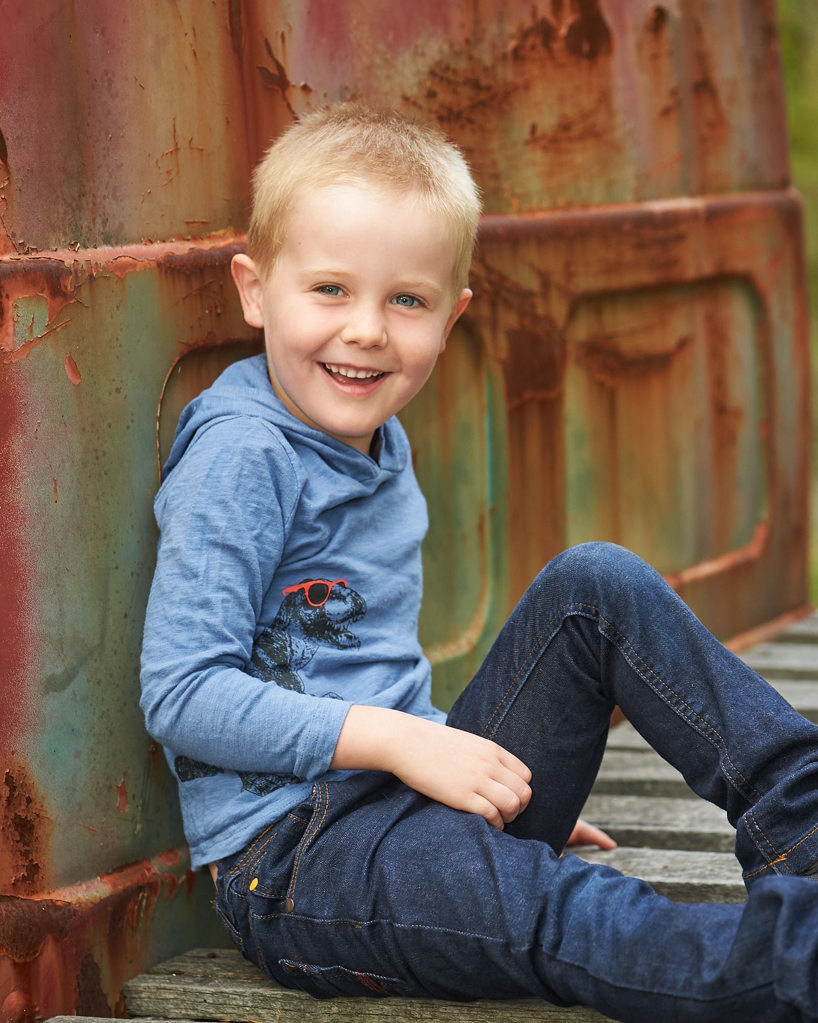 Smiling boy in a blue hoodie sits on timber boards against a rusty vintage truck