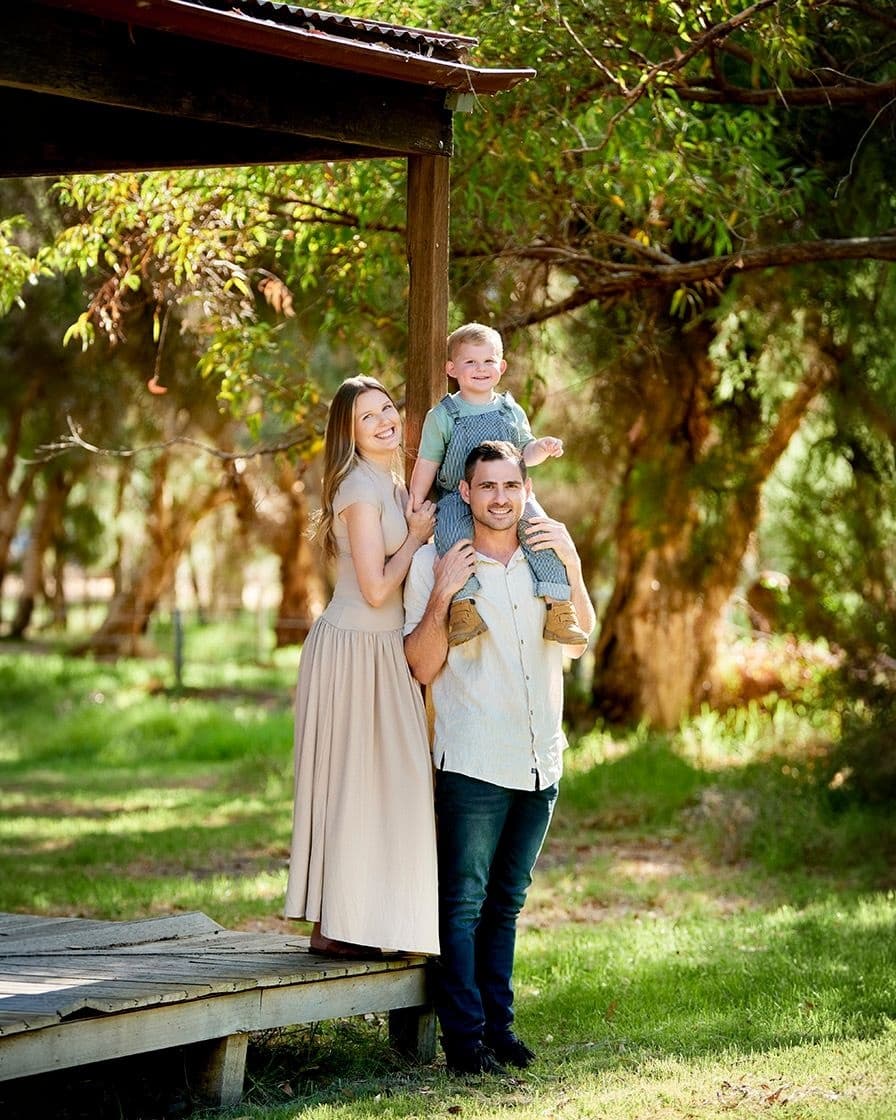 A mum smiling beside her husband, their toddler on his shoulders, under the trees
