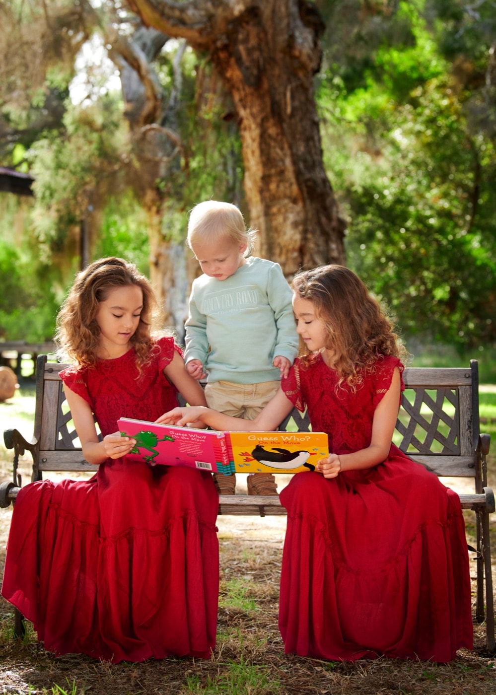 Two sisters in red dresses reading with their little brother on a garden bench