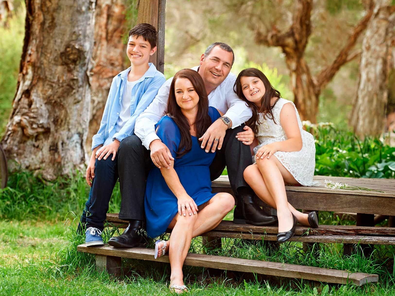 A family of four sitting together on timber steps under the paperbarks