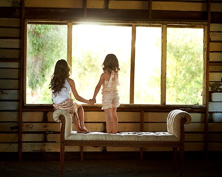 Two girls holding hands on a tufted bench, gazing out of a sunlit cottage window