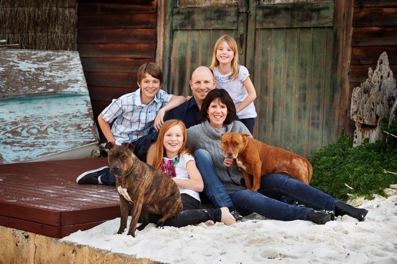 Family of five with two dogs sitting on white sand in front of the boat shed