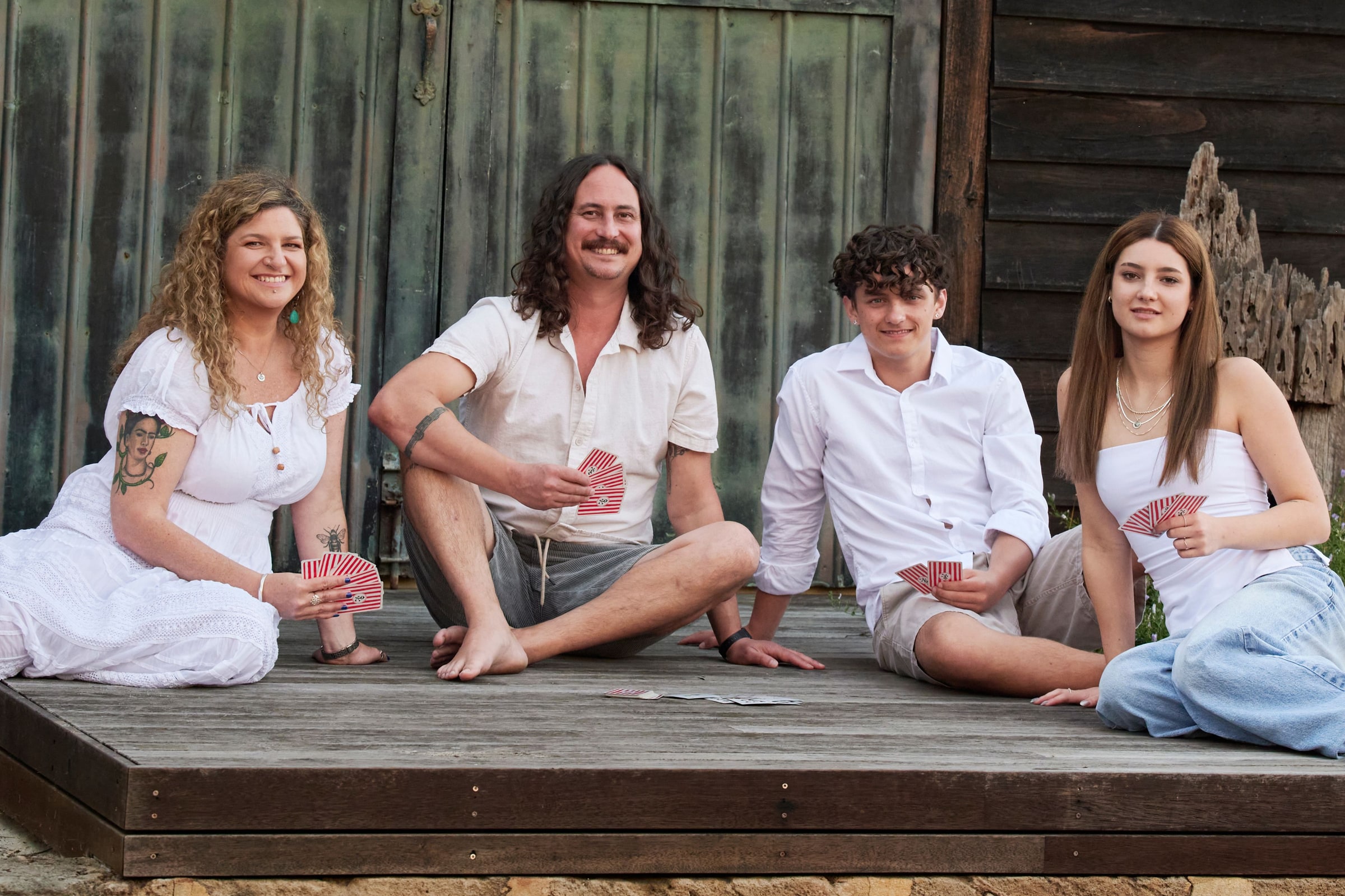 Parents and two teenagers sit in a row on the boat shed deck holding playing cards