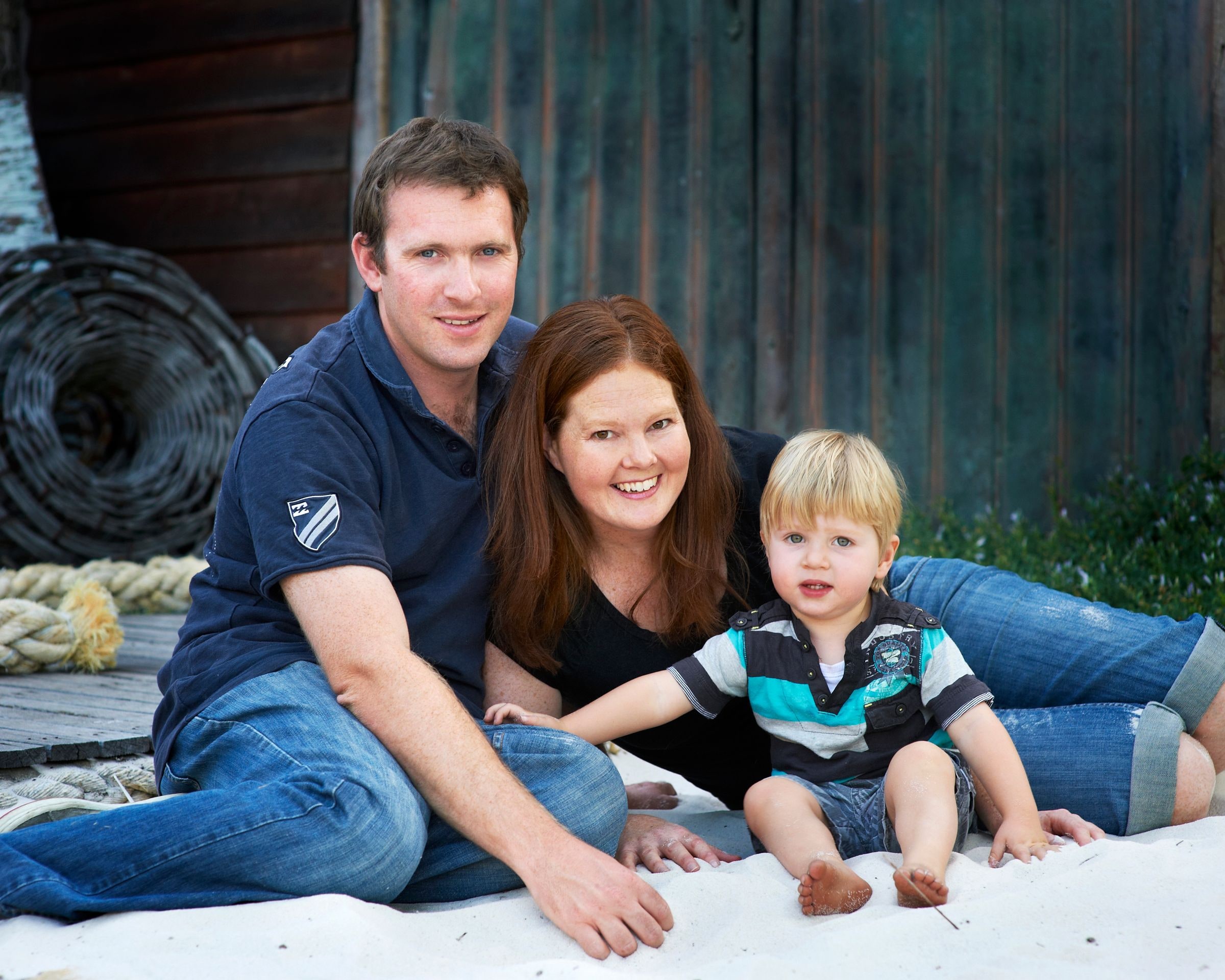 Parents and their toddler sitting on the sand by the boat shed doors