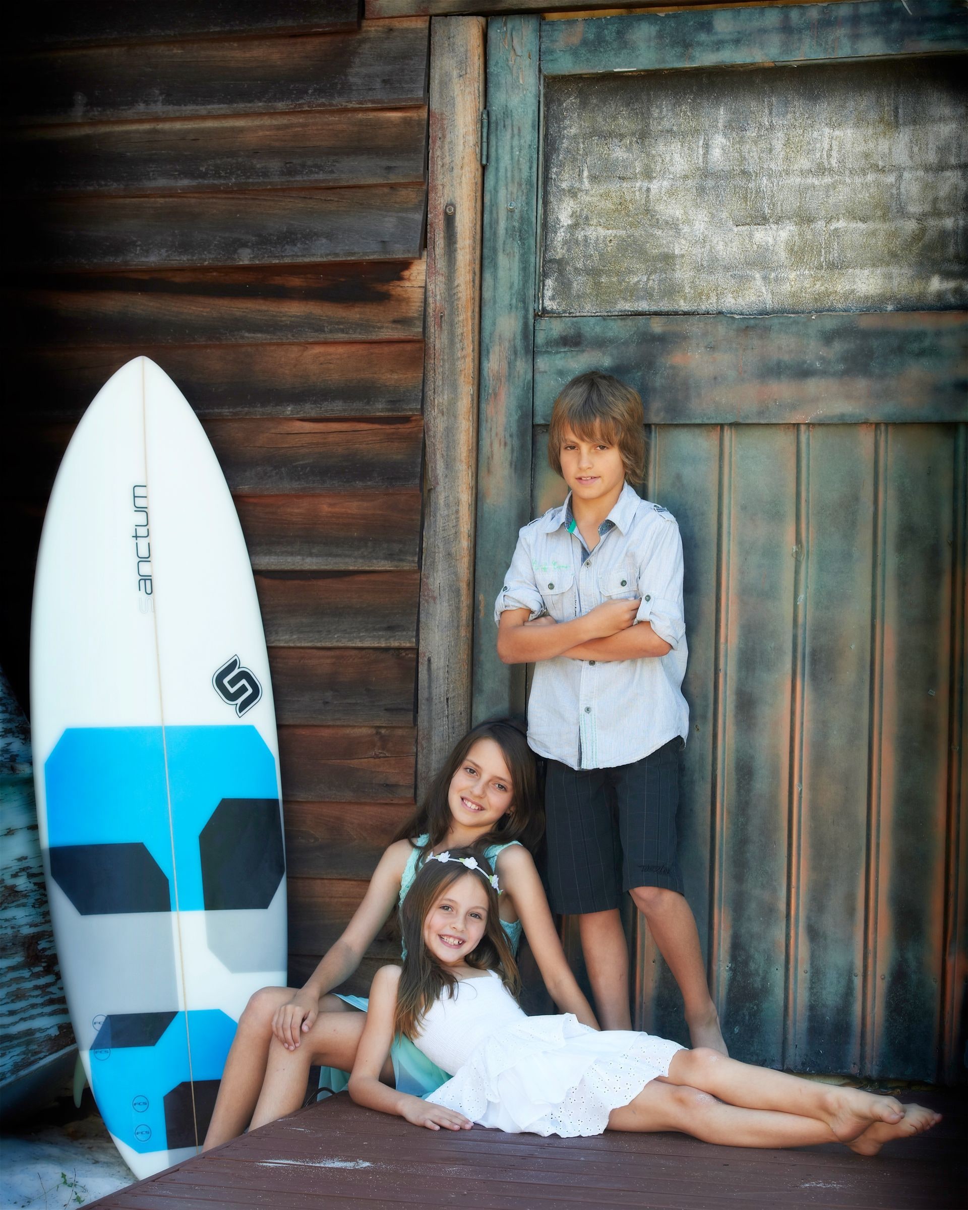 Three children with a surfboard against the old boat shed doors