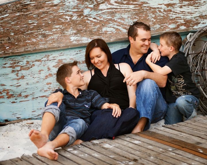 Parents and two boys sit close on a timber deck against a weathered blue boat