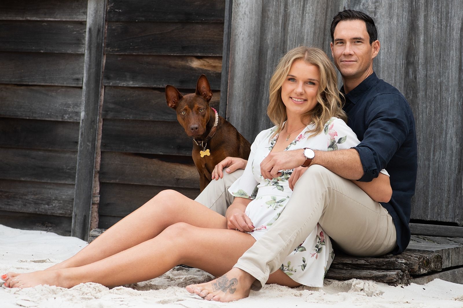 A couple sitting on the sand with their dog beside the boat shed