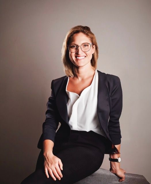 Woman in glasses and a dark blazer smiling while seated against a grey studio backdrop