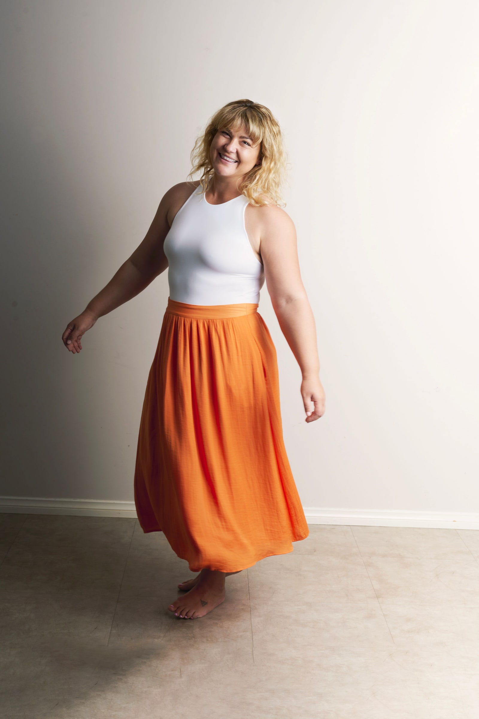 Barefoot woman in a white top and orange skirt smiles mid-twirl in the studio