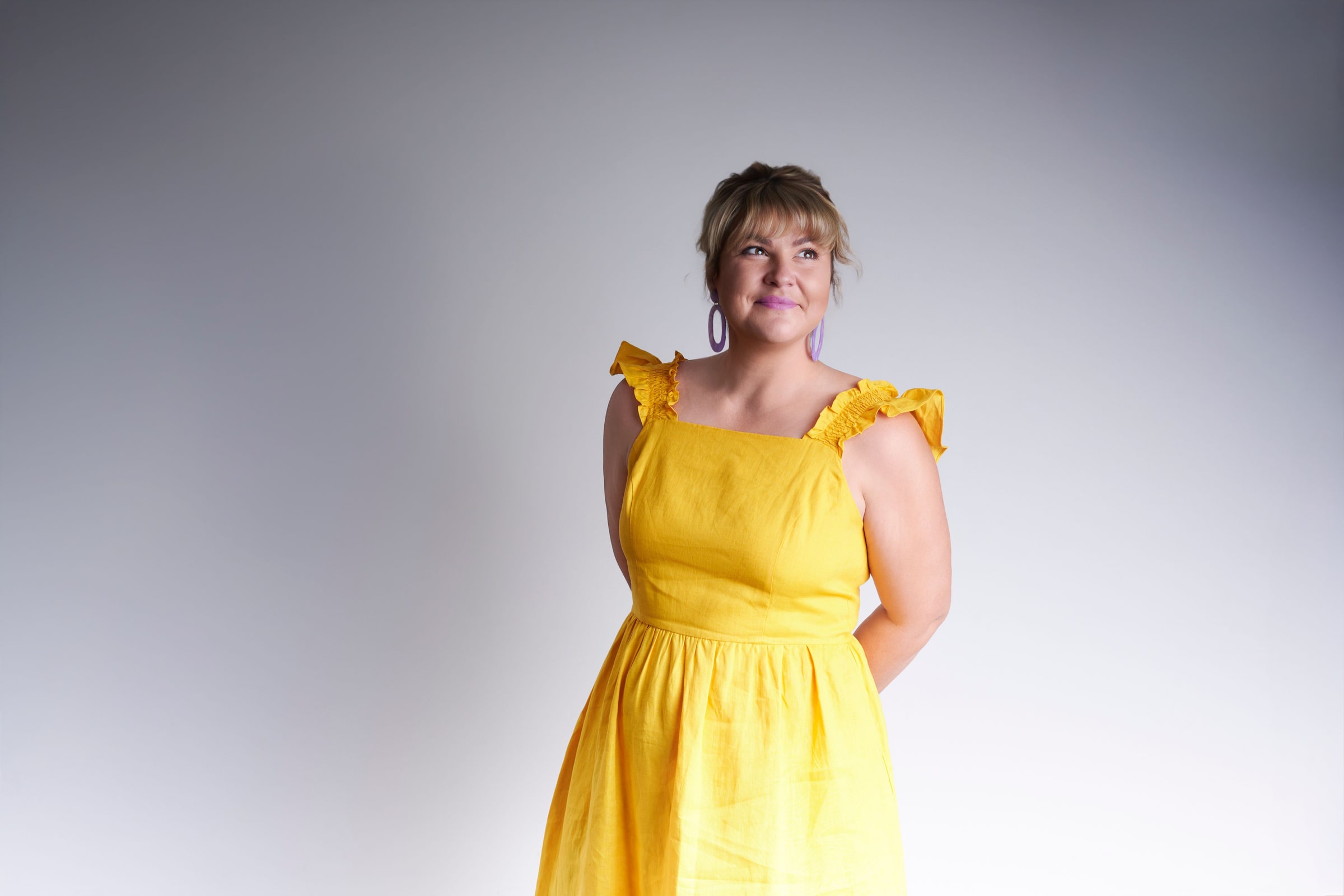 Woman in a yellow dress with ruffled straps smiles and looks upward against a grey backdrop