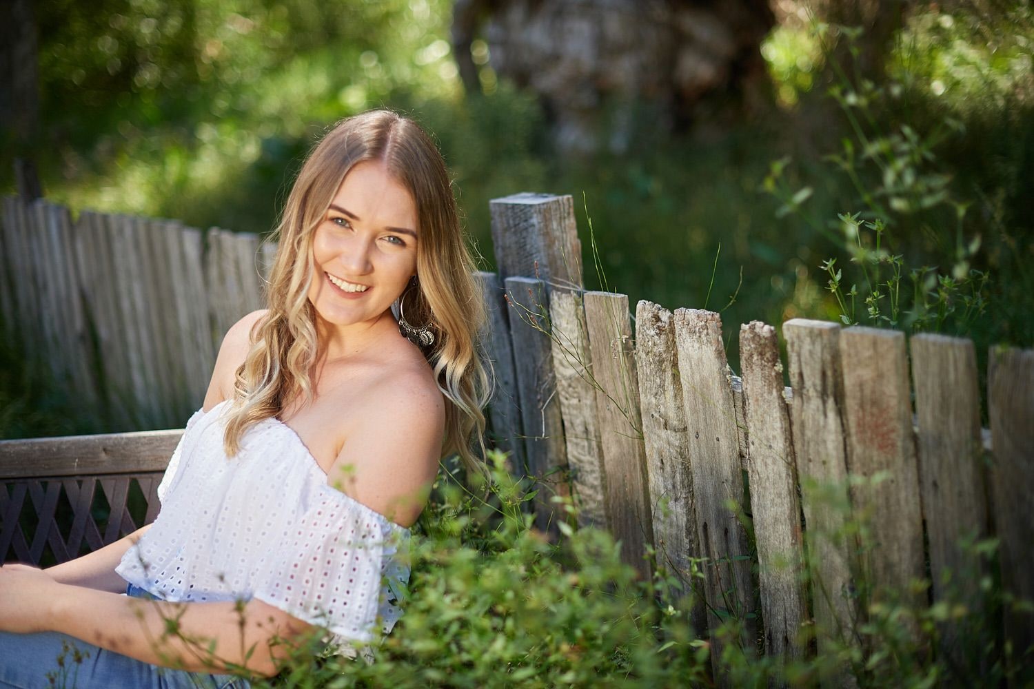 Young woman in a white off-shoulder top smiling beside a weathered timber picket fence