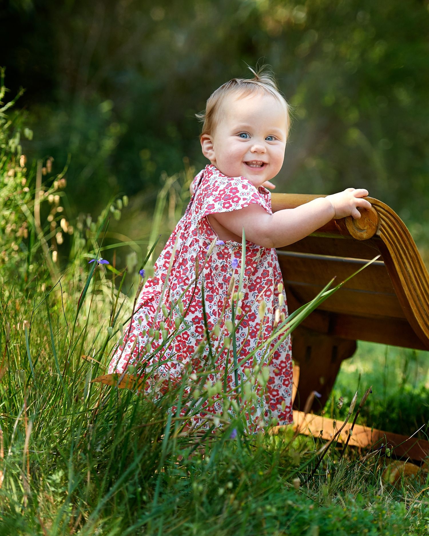 A baby girl in a floral dress standing beside a timber chair in the grass