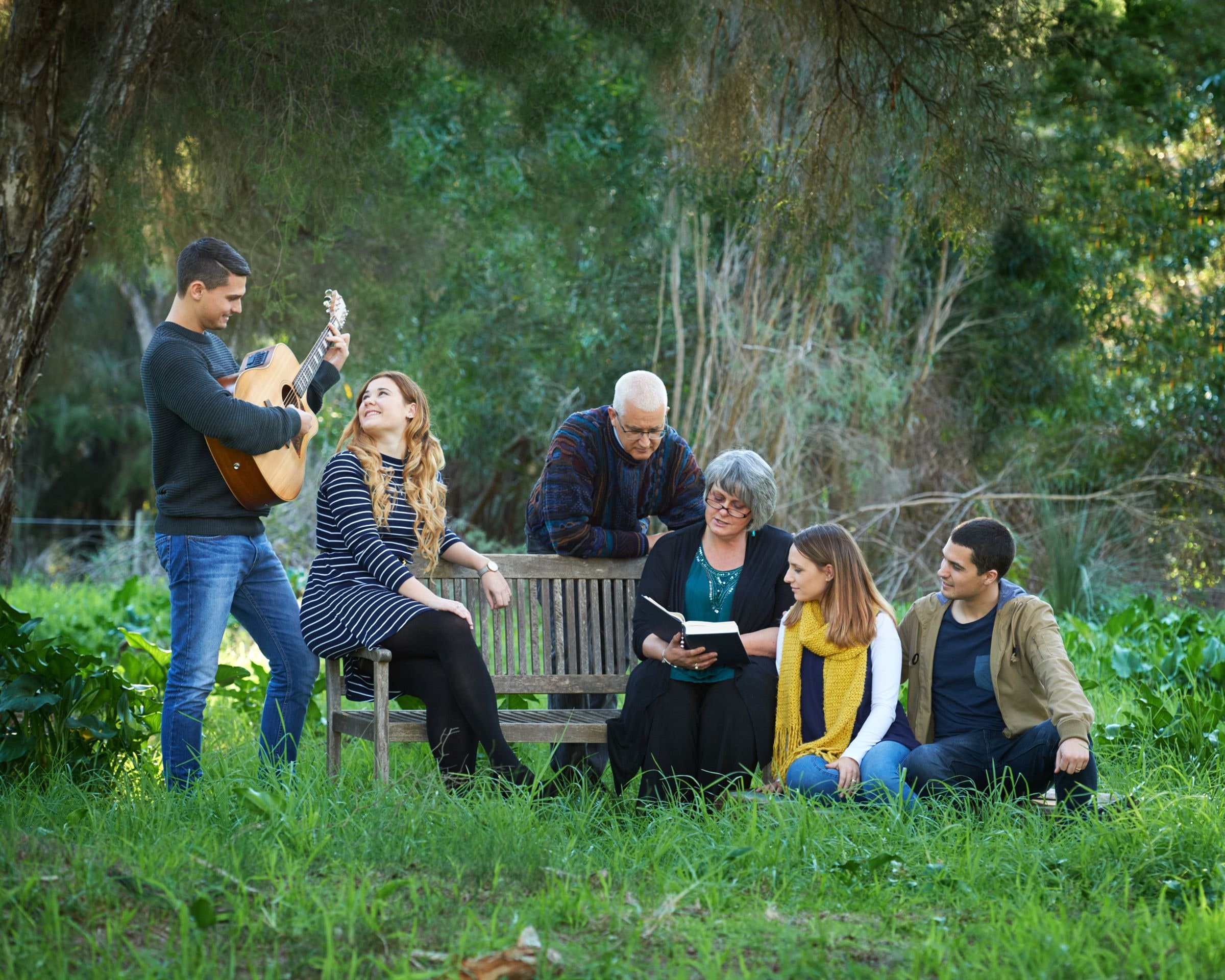 A family of seven gathered around a timber bench in the bushland, one with a guitar