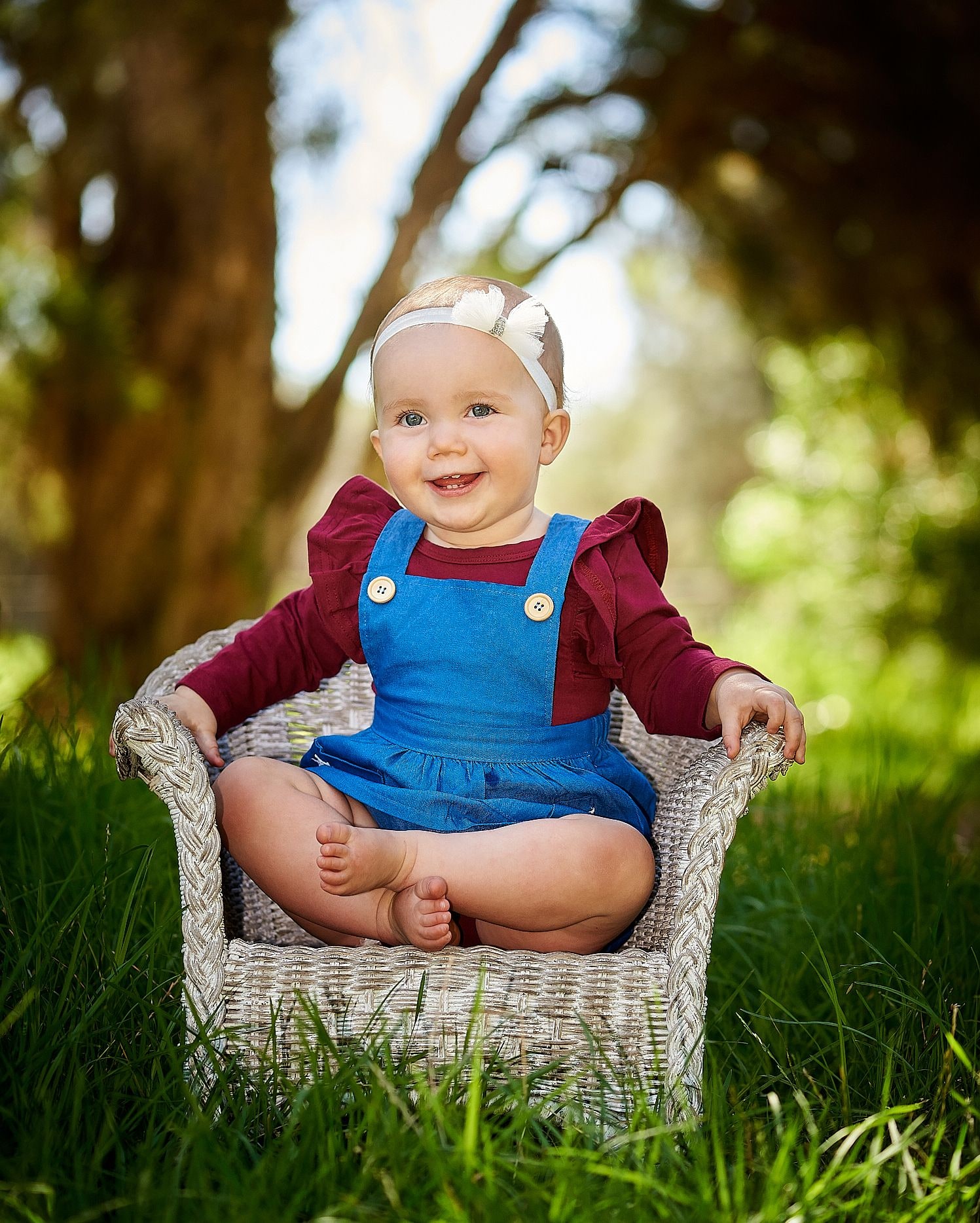 Smiling baby in a denim pinafore sits cross-legged in a wicker chair on bushland grass