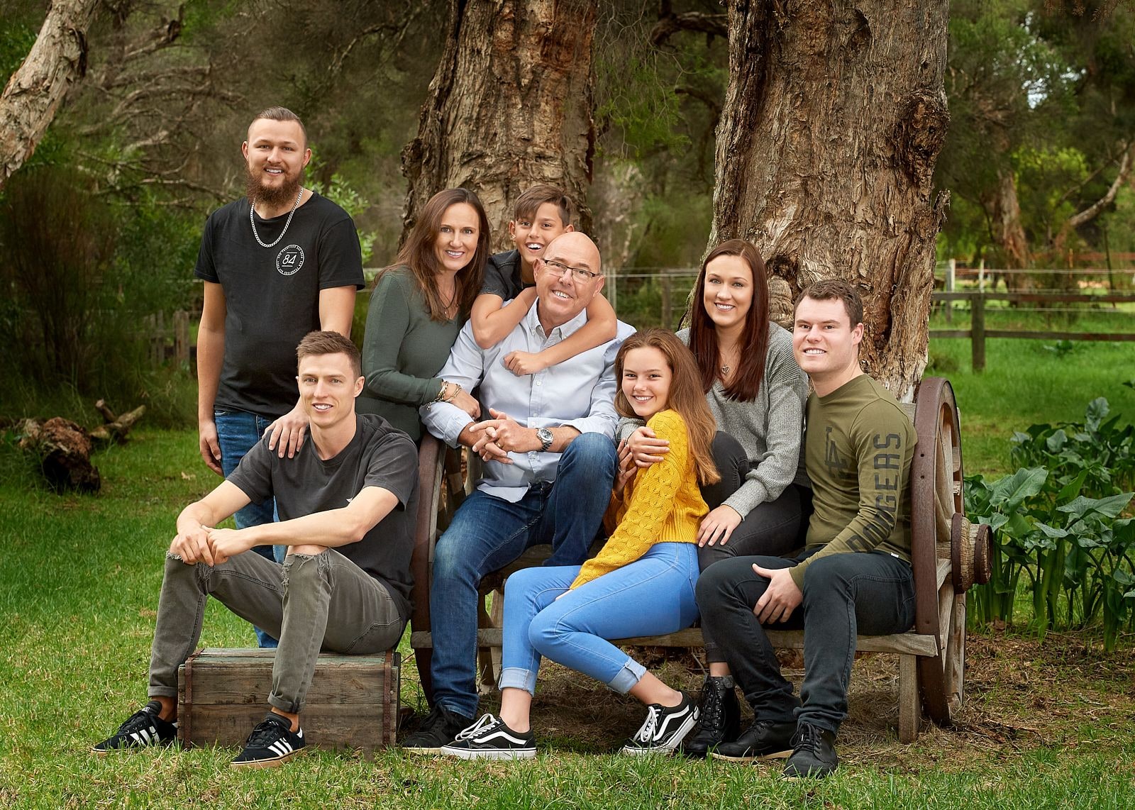 Large family of eight gathered on a wagon-wheel bench and crate beneath tall bushland trees