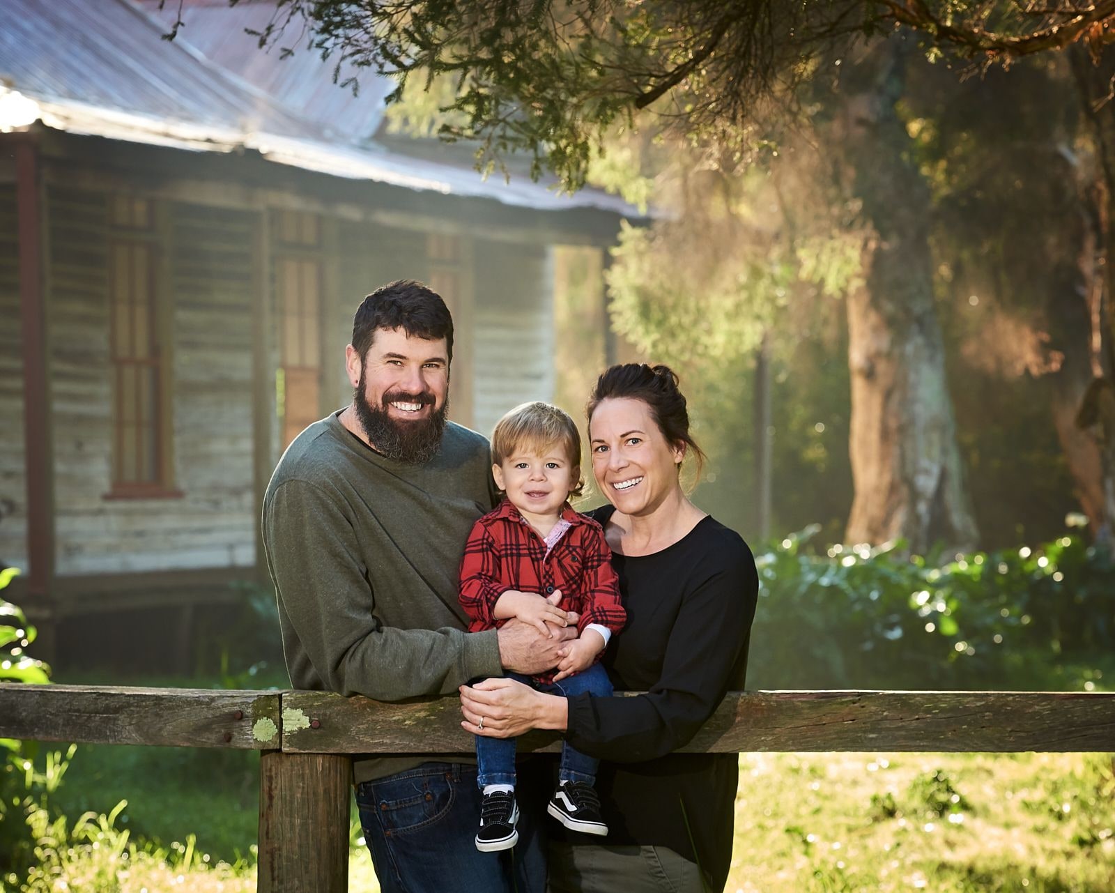Parents holding their toddler son at a timber rail fence in front of an old cottage