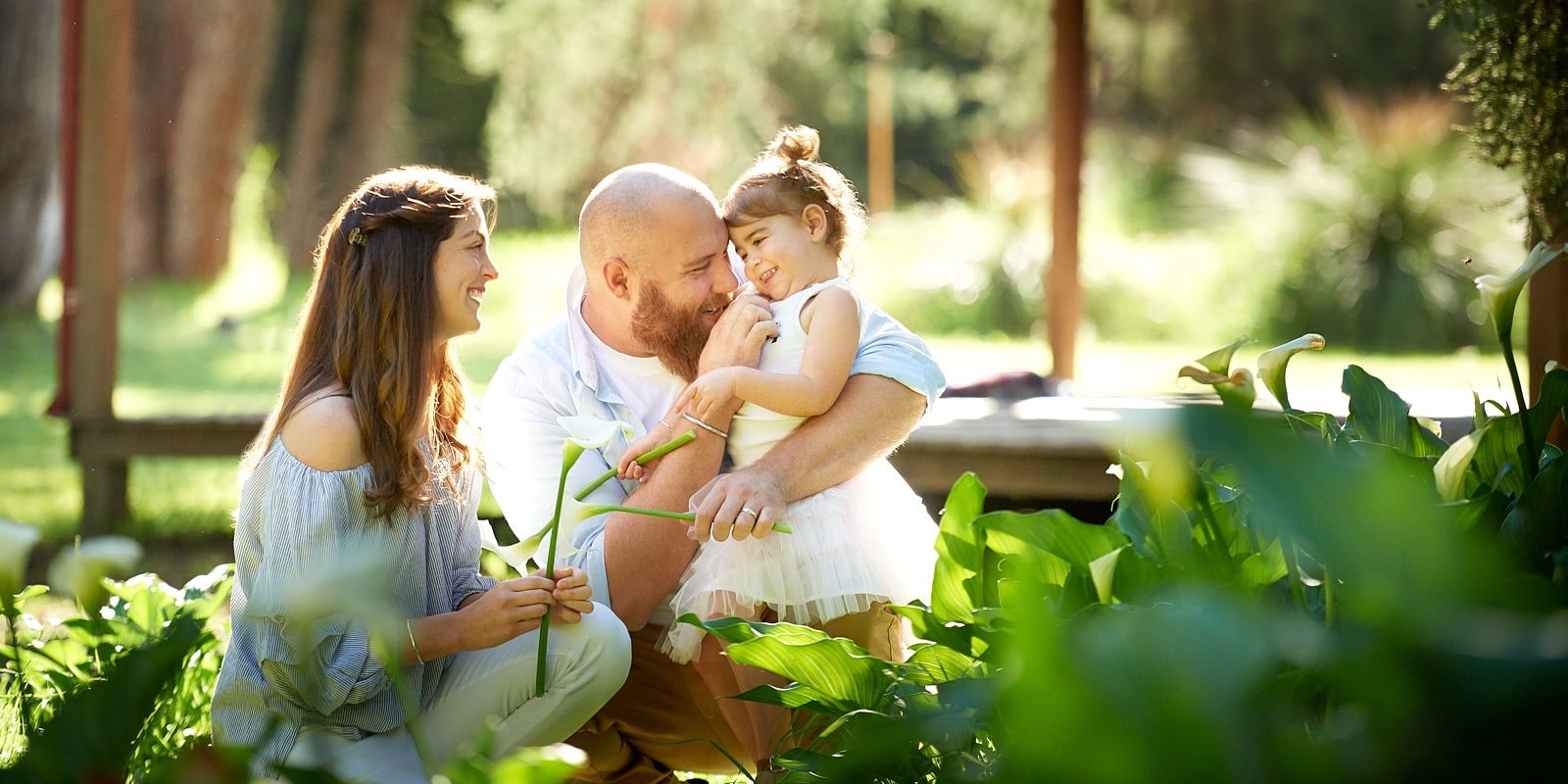 Father cuddling his toddler daughter in a white tutu beside mother among arum lilies