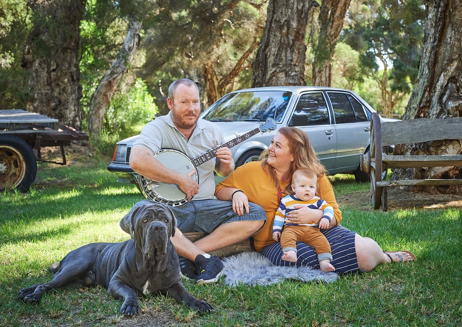 Father playing a banjo beside mother and baby, a large grey dog lying in front