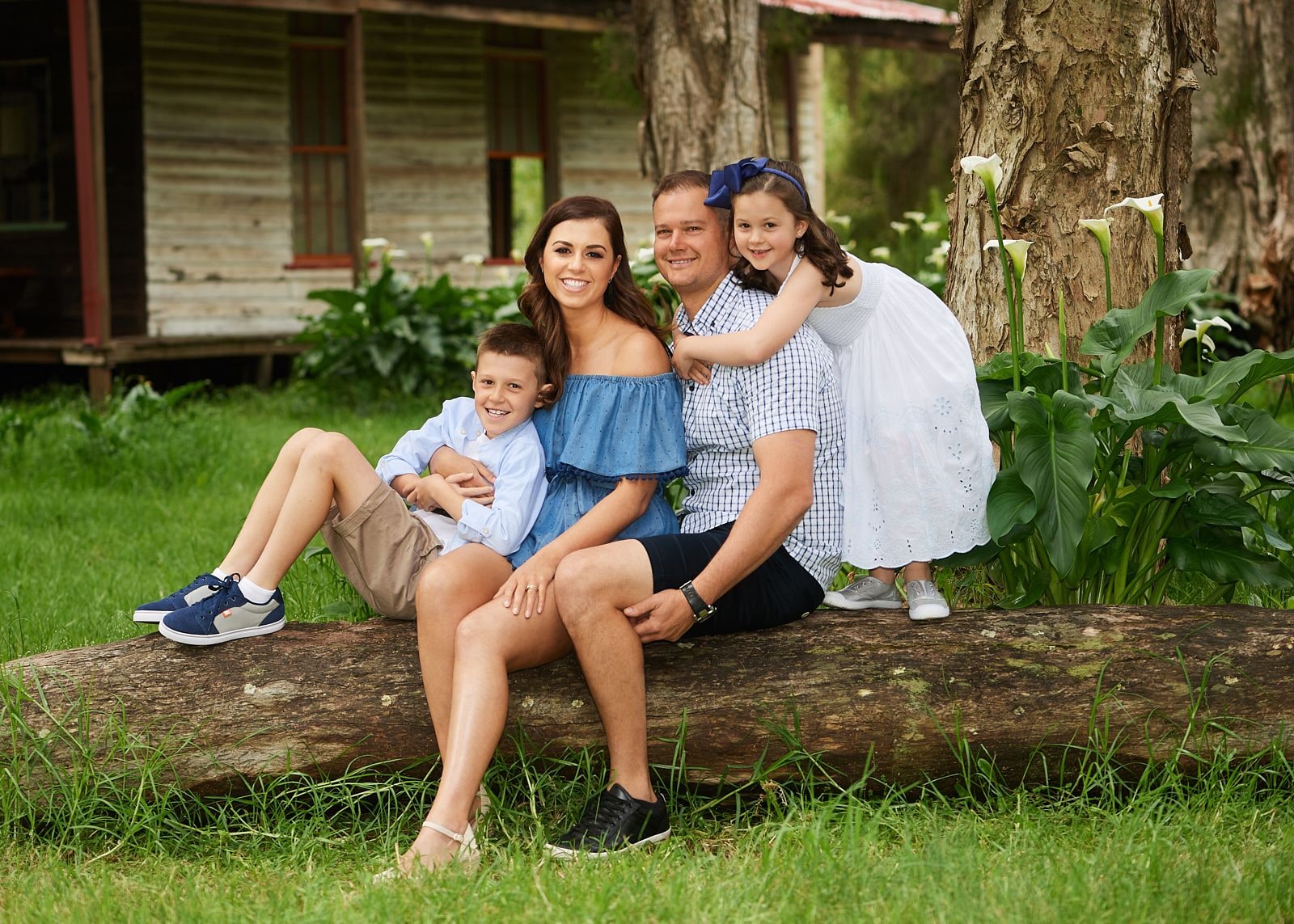 Family of four on a fallen log, daughter hugging her dad from behind, near arum lilies
