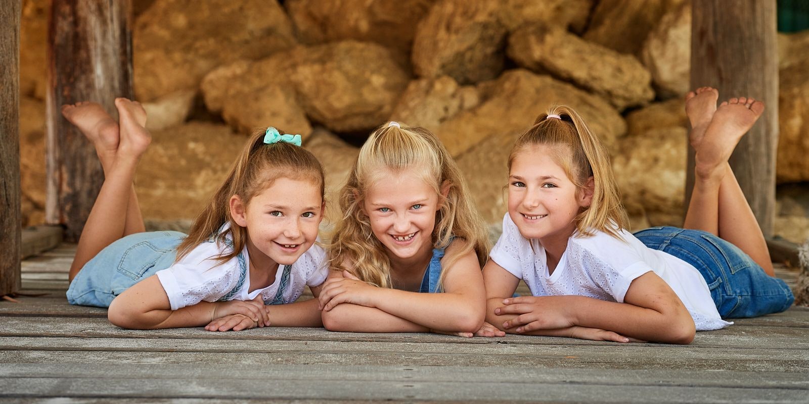 Three girls lie on their tummies on a timber jetty deck with feet kicked up