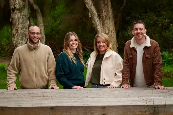 Four adults in cream, tan, forest green and brown standing at the timber bench at Premier Studio