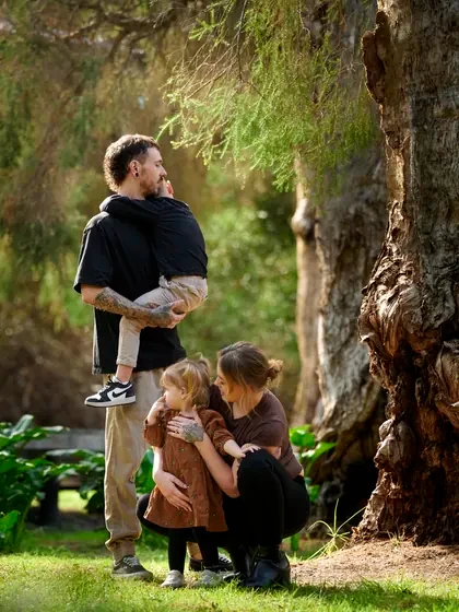 Family in black, brown and tan gathered beneath the paperbark trees at Premier Studio
