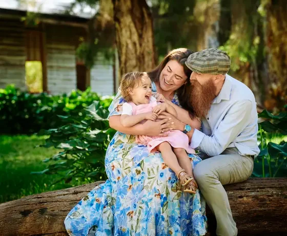 Mum in a blue floral dress with dad in tan and baby in pink on a log at Premier Studio