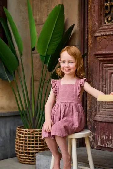 Little girl in a dusty rose pinafore dress seated by the aged timber doors at Premier Studio