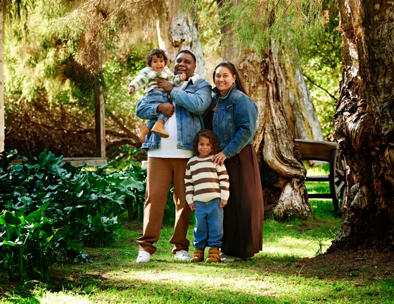 Family of four in denim jackets, cream stripes and tan standing by the paperbark at Premier Studio